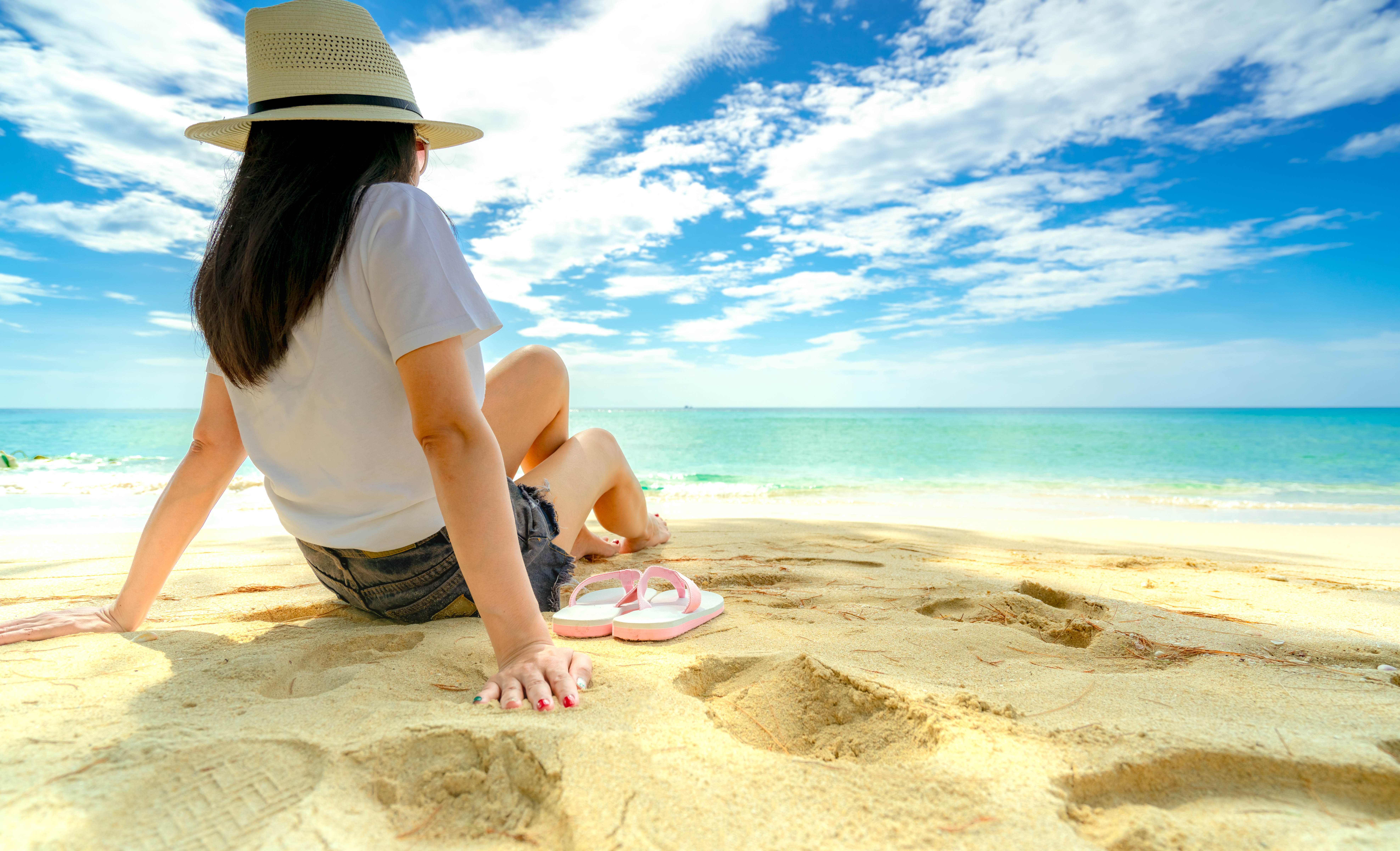 Eine junge Frau sitzt mit einem Sonnenhut am Strand und betrachtet das Meer